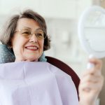 Uśmiechnięta starsza pacjentka oglądająca uzębienie w lusterku po leczeniu protetycznym w Klinice Biały Kamień w Nowym Mieście nad Pilicą