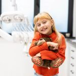 Uśmiechnięta dziewczynka z pluszowym misiem w gabinecie stomatologii dziecięcej Klinika Biały Kamień w Nowym Mieście nad Pilicą